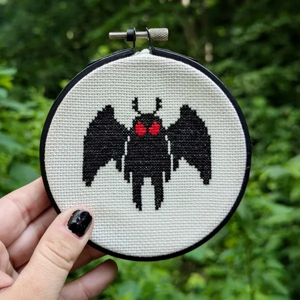 Mini Mothman cross stitch on cream fabric held in a hand